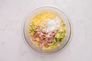 A glass mixing bowl containing grated cheese, flour, sliced ham, shredded zucchini, and beaten eggs on a white marble surface, ready to be mixed together.