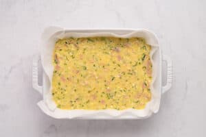 A white baking dish lined with parchment paper, filled with an uncooked mixture of eggs, herbs, and diced ham or vegetables, ready to be baked.