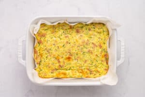 A baked zucchini slice with a golden top sits in a white rectangular dish lined with parchment paper, on a light marble surface.