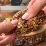 Close-up of hands breaking apart a pastry filled with chopped pistachios. The gooey filling is being stretched, and the wooden cutting board below has scattered crumbs. A blurred teapot is visible in the background.