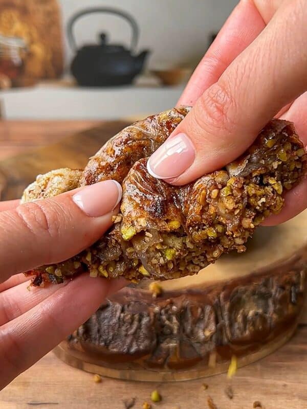 Close-up of hands breaking apart a pastry filled with chopped pistachios. The gooey filling is being stretched, and the wooden cutting board below has scattered crumbs. A blurred teapot is visible in the background.