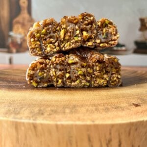 Two nut-filled date rolls stacked on a wooden surface, showing a dense, textured filling of chopped pistachios and nuts inside a chewy brown outer layer. The background is softly blurred.