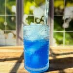A textured glass filled with blue liquid and ice sits on a wooden surface in sunlight, with a clear straw and a blurred window showing green foliage in the background.