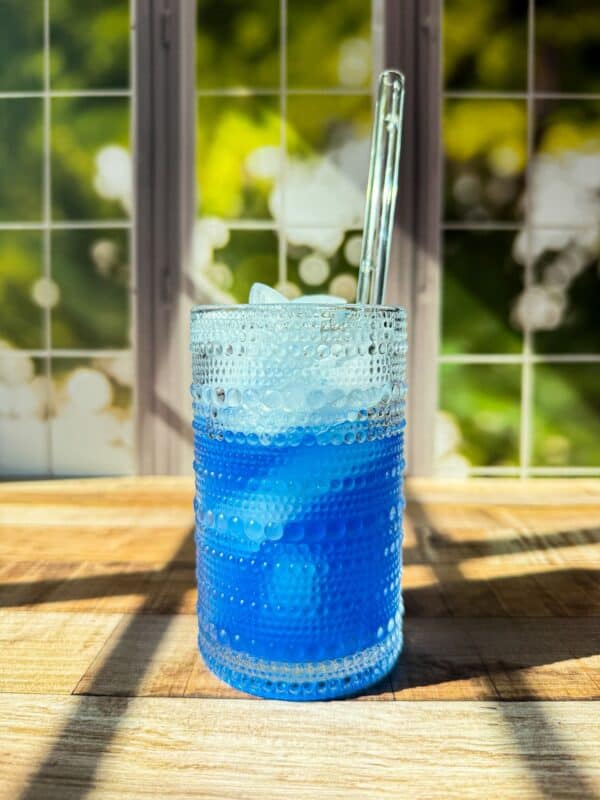 A textured glass filled with blue liquid and ice sits on a wooden surface in sunlight, with a clear straw and a blurred window showing green foliage in the background.