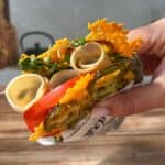 A hand holding a sandwich with crispy cheese, pickles, tomato, greens, and rolled slices of deli meat, wrapped partially in paper, over a wooden table and gray background.
