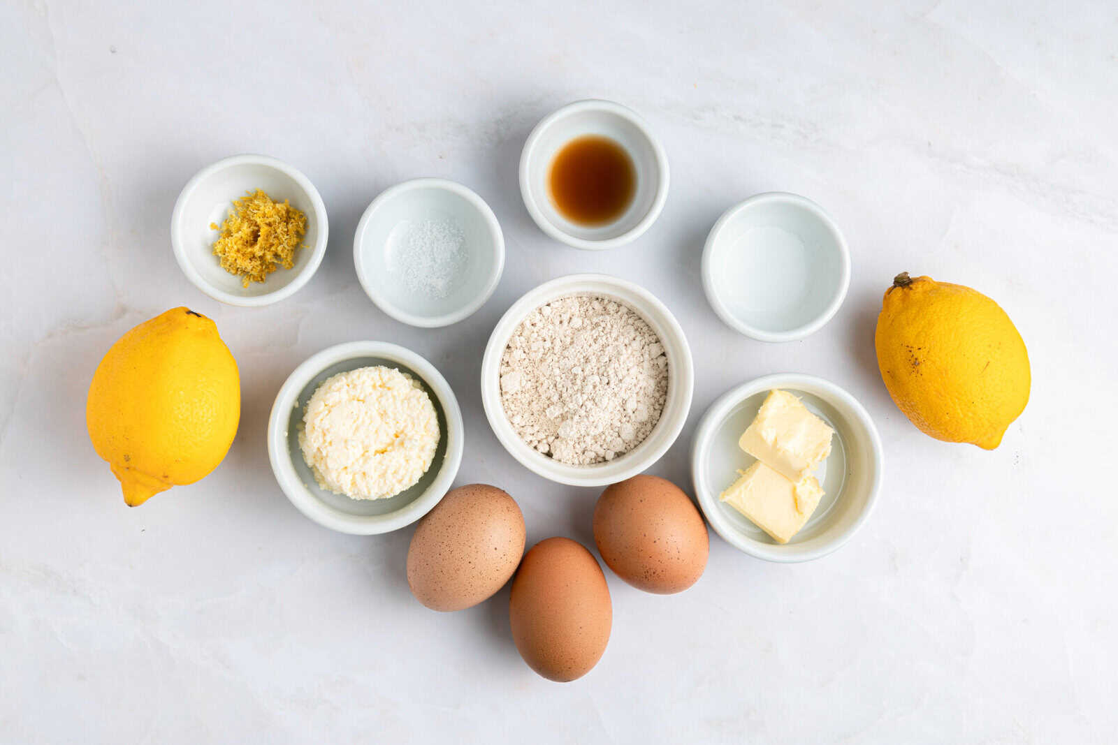 A flat lay of baking ingredients on a white surface, including two whole lemons, three brown eggs, flour, butter, ricotta cheese, lemon zest, vanilla extract, baking powder, and two small bowls with clear liquids.