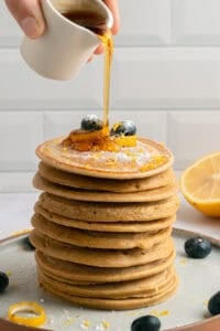 A hand pours syrup from a small white pitcher onto a tall stack of pancakes topped with blueberries and powdered sugar, with a lemon half and more blueberries on the plate.