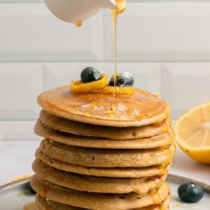 A hand pours syrup from a small white pitcher onto a tall stack of pancakes topped with blueberries and lemon zest, with syrup dripping down the sides. A halved lemon and scattered blueberries sit nearby.