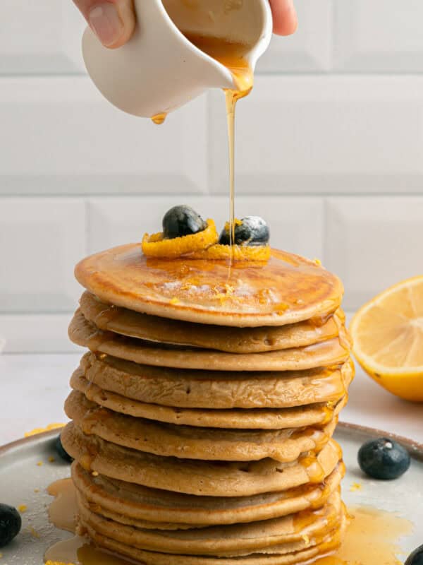 A hand pours syrup from a small white pitcher onto a tall stack of pancakes topped with blueberries and lemon zest, with syrup dripping down the sides. A halved lemon and scattered blueberries sit nearby.