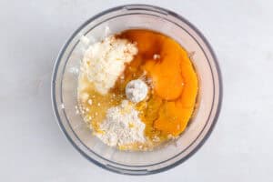 A food processor bowl containing eggs, flour, ricotta cheese, and other baking ingredients, viewed from above on a light countertop.
