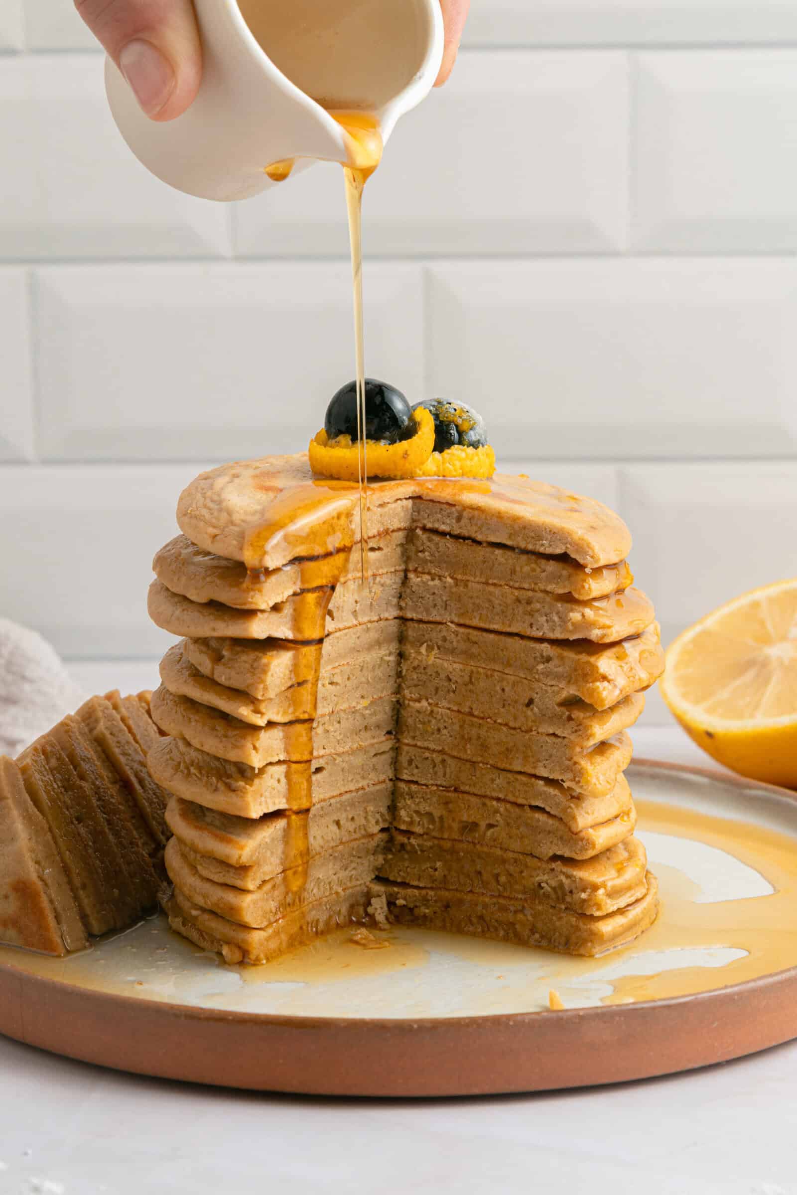 A stack of sliced pancakes topped with blueberries and an orange slice is drizzled with syrup from a small white pitcher, with a lemon half in the background.
