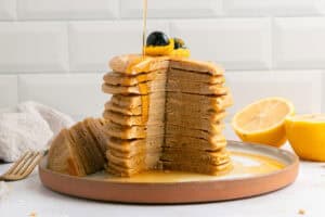 A tall stack of pancakes with a large wedge cut out is topped with blueberries and zest, with syrup being poured over. A halved lemon sits beside the plate on a white countertop.