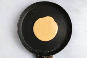 A pancake is cooking in a black nonstick frying pan, with batter spread in a round shape on the pan’s surface. The background is a light-colored countertop.