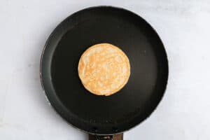 A single pancake cooking in the center of a black frying pan on a light-colored countertop.