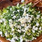 A wooden bowl filled with chopped lettuce, fresh herbs, and crumbled white cheese, with wooden salad servers resting on the edge. The salad sits on a wooden surface.