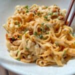 A bowl of wide, flat noodles tossed in a creamy, spicy sauce, garnished with chopped chives. Chopsticks rest on the edge of the bowl. The noodles are served on a light-colored, speckled ceramic dish.