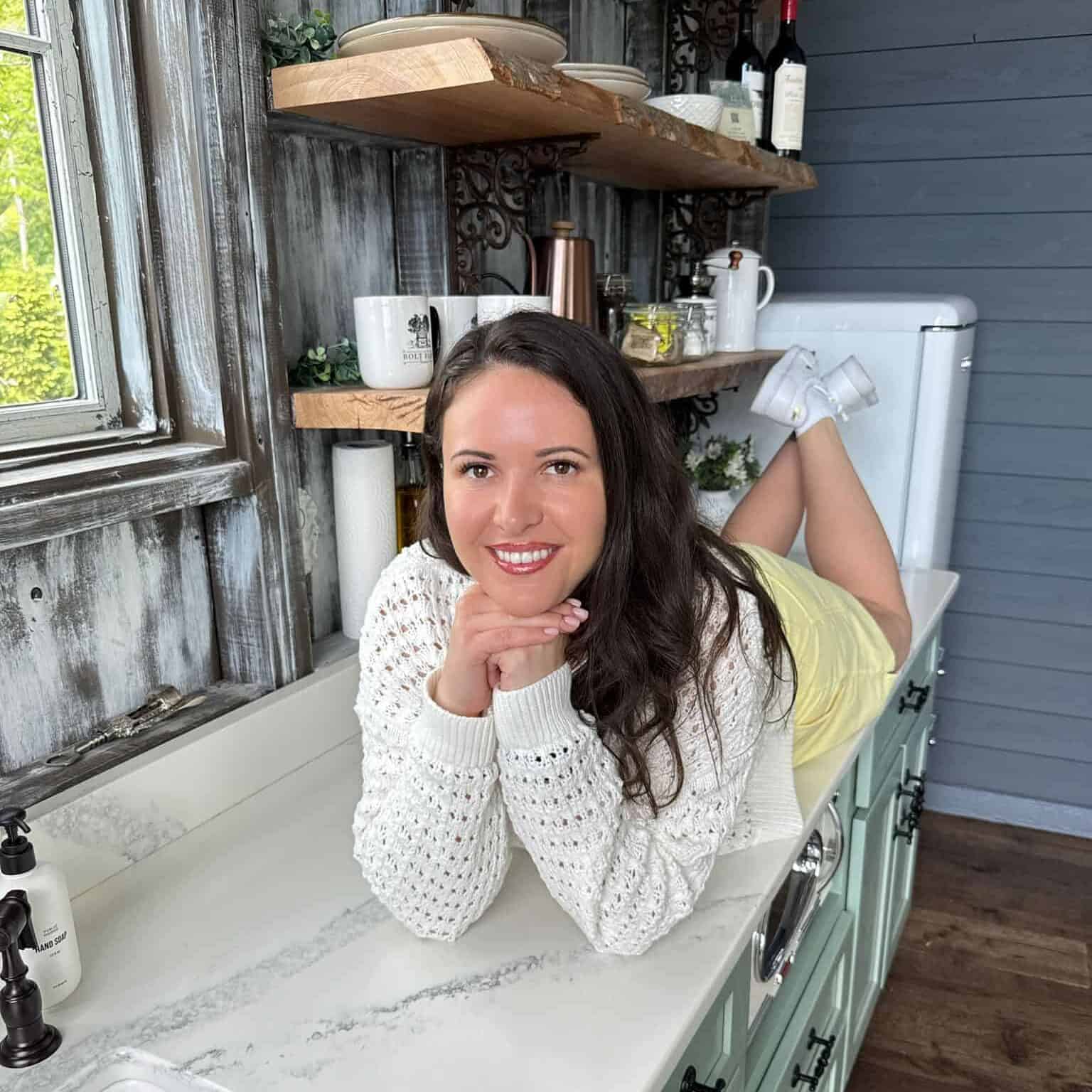 The Modern Nonna with long brown hair, wearing a white knit sweater and light yellow skirt, smiles while lying on her stomach on a kitchen counter with her legs bent and hands under her chin. Wooden shelves and kitchen items are in the background.