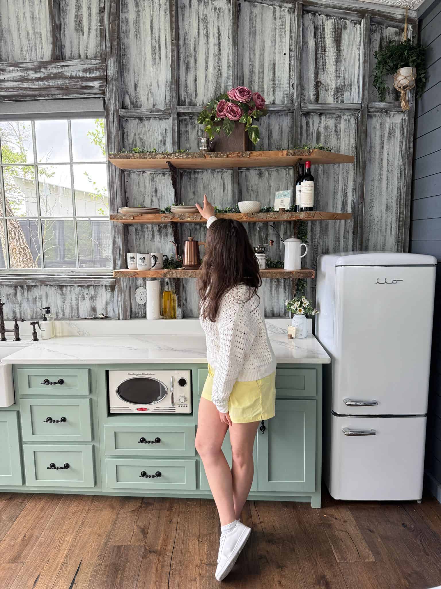 The Modern Nonna (Sneji) in a white sweater and yellow shorts stands in a rustic kitchen, pointing up at a shelf with pink flowers. The kitchen has mint green cabinets, open wooden shelves, and a vintage white refrigerator.