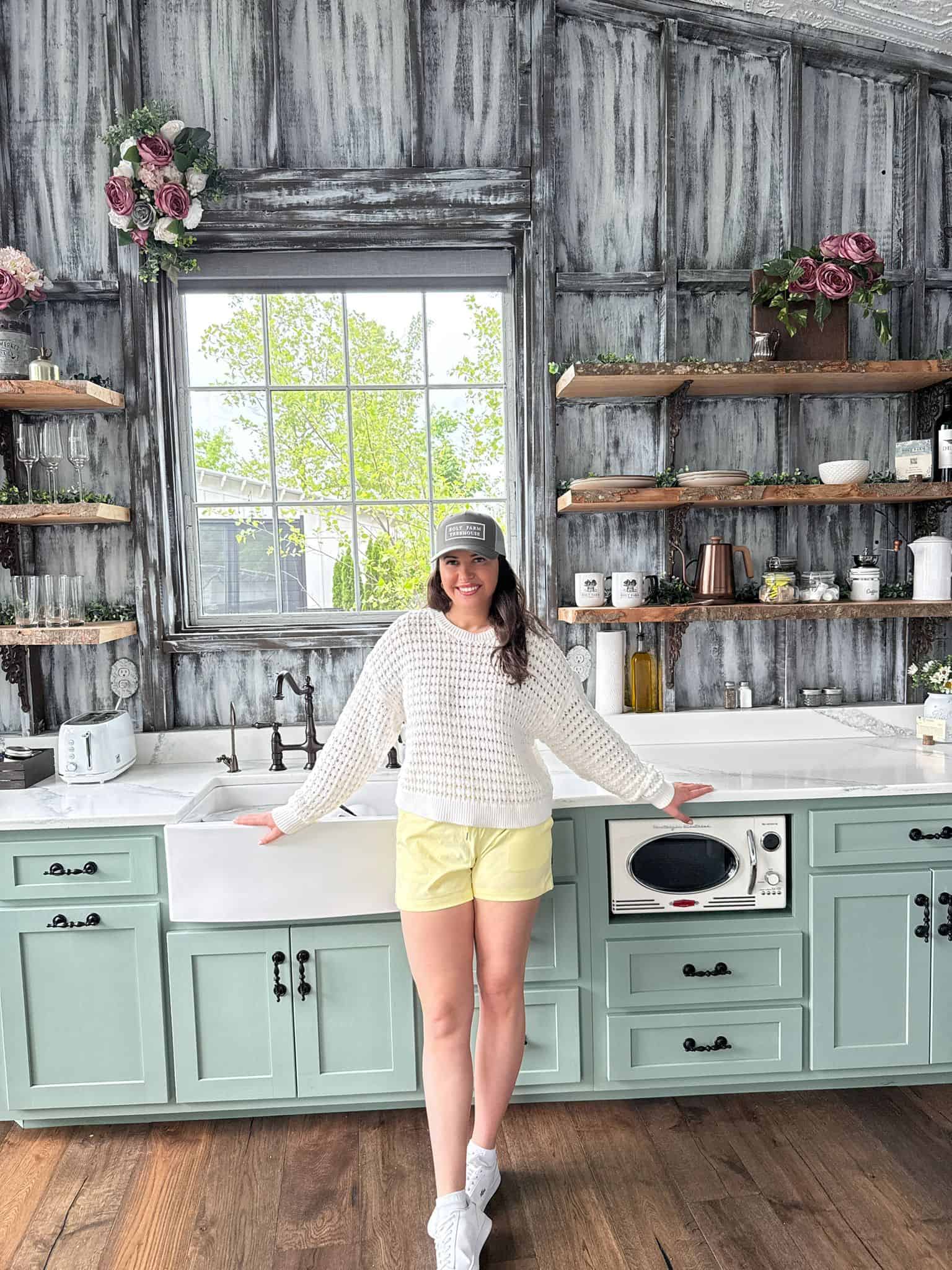 A woman in a white sweater, yellow shorts, and a gray cap smiles while standing in a vintage-style kitchen with mint green cabinets, open shelves, and a large window behind her.
