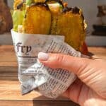 A hand holding a paper-wrapped bundle of deep-fried vegetable slices, including zucchini and possibly green beans, with a rustic wooden table and blurred background in sunlight.