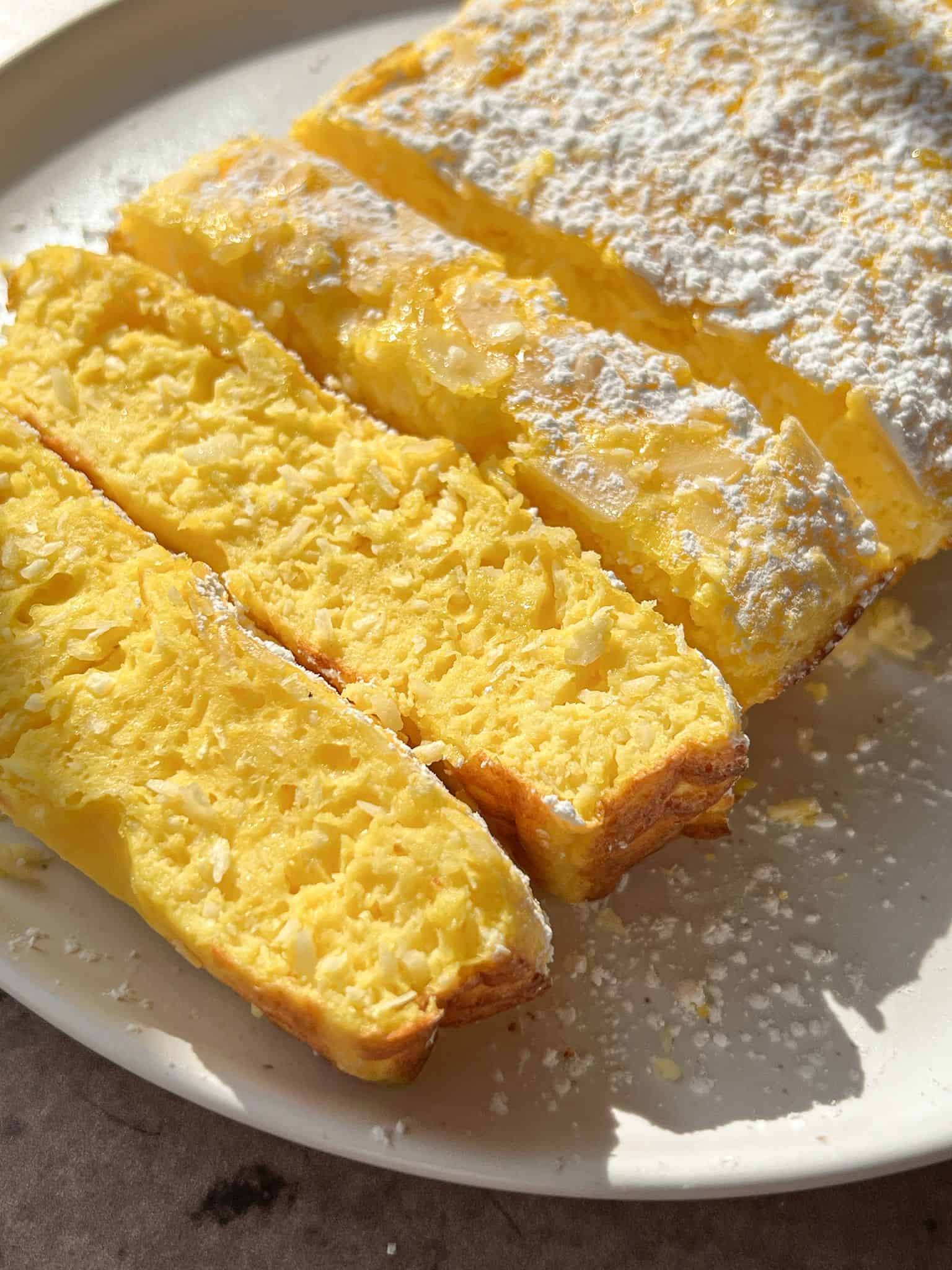 Close-up of thick, golden-yellow slices of baked custard or egg-based dessert dusted with powdered sugar, arranged on a white plate in sunlight.