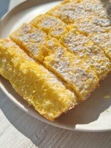 A close-up of a sliced, yellow, cake-like dessert dusted with powdered sugar on a beige plate. The texture appears moist and fluffy, with visible coconut flakes inside. Sunlight highlights the surface.