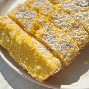 A close-up of a sliced, yellow, cake-like dessert dusted with powdered sugar on a beige plate. The texture appears moist and fluffy, with visible coconut flakes inside. Sunlight highlights the surface.