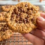 A hand holds an oatmeal cookie topped with chocolate chips over a cooling rack with more similar cookies. Additional baked goods are blurred in the background.