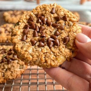 A hand holds an oatmeal cookie topped with chocolate chips over a cooling rack with more similar cookies. Additional baked goods are blurred in the background.