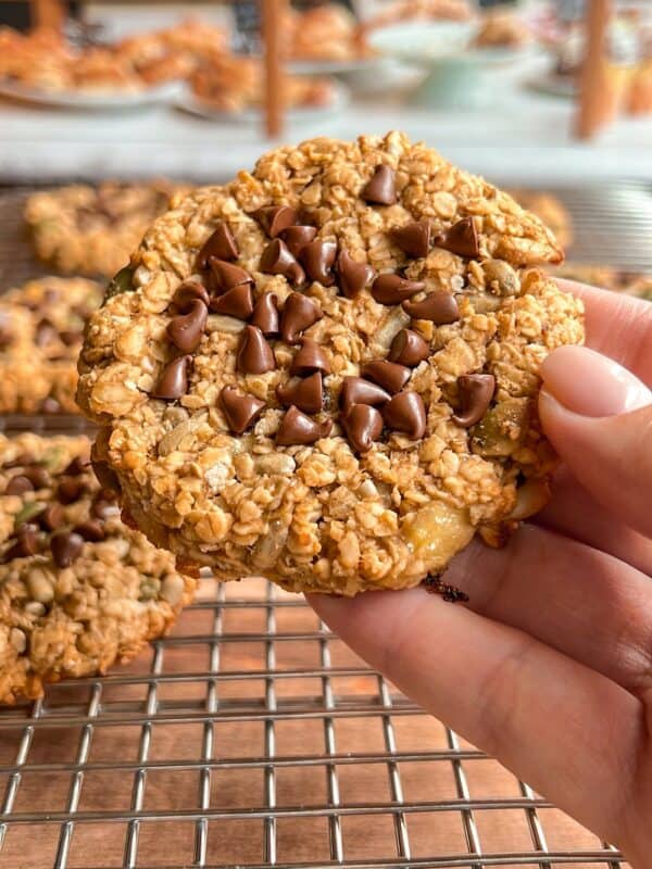 A hand holds an oatmeal cookie topped with chocolate chips over a cooling rack with more similar cookies. Additional baked goods are blurred in the background.