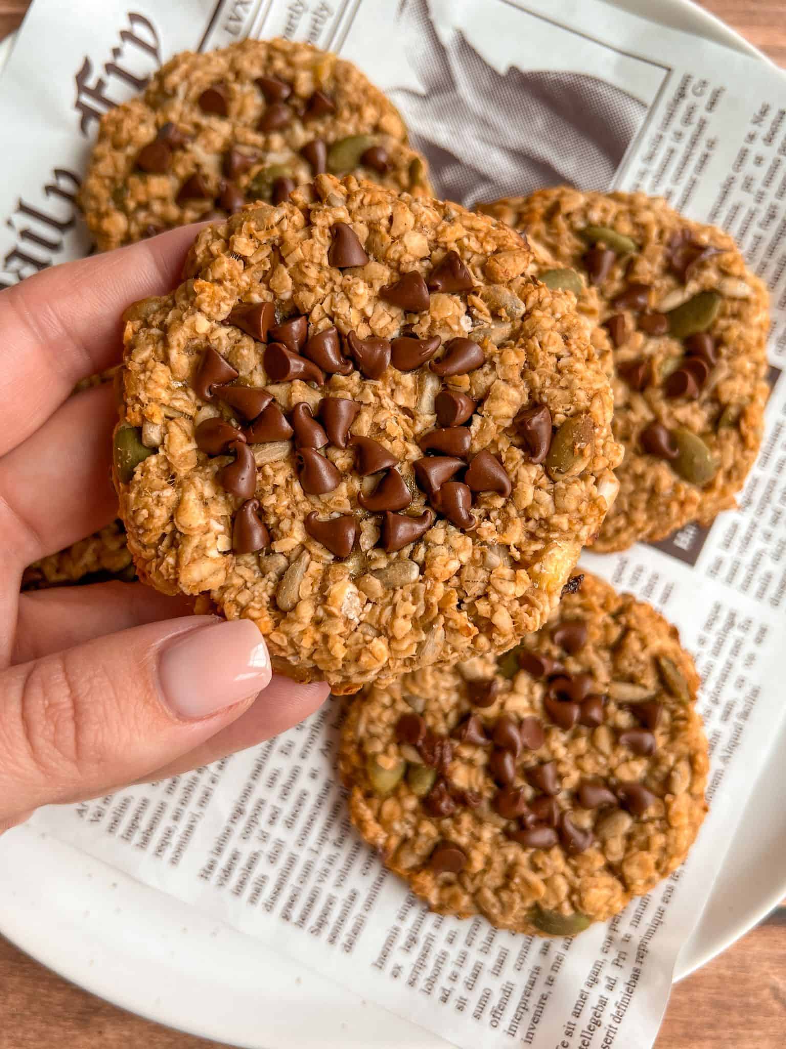 A hand holding an oatmeal cookie topped with chocolate chips and seeds, with more cookies on a plate lined with newspaper-style paper in the background.