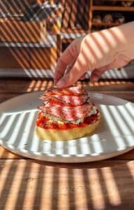 A hand places slices of salami on top of bread with red sauce and cheese, on a white plate. Sunlight streams through blinds, casting striped shadows over the food and table.