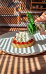 A hand sprinkles dried herbs from a green spice container onto a slice of bread topped with tomato sauce and cheese, placed on a white plate with sunlight and shadows across the scene.