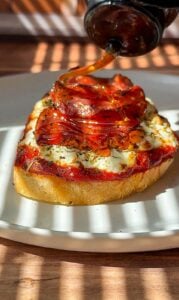 A slice of bread topped with cheese, tomato sauce, and pepperoni is served on a plate while honey is being drizzled over it. Sunlight and shadows create a striped pattern across the scene.