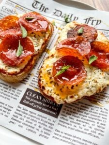 Two slices of toasted bread topped with creamy cheese, pepperoni slices, and small green herb leaves, served on a newspaper-themed paper.