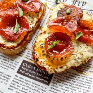 Two slices of toasted bread topped with creamy cheese, pepperoni slices, and small green herb leaves, served on a newspaper-themed paper.
