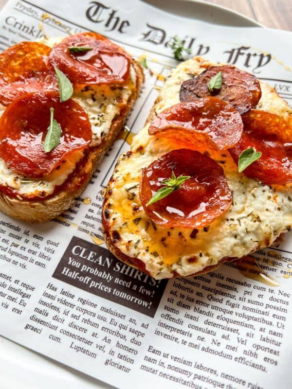 Two slices of toasted bread topped with creamy cheese, pepperoni slices, and small green herb leaves, served on a newspaper-themed paper.