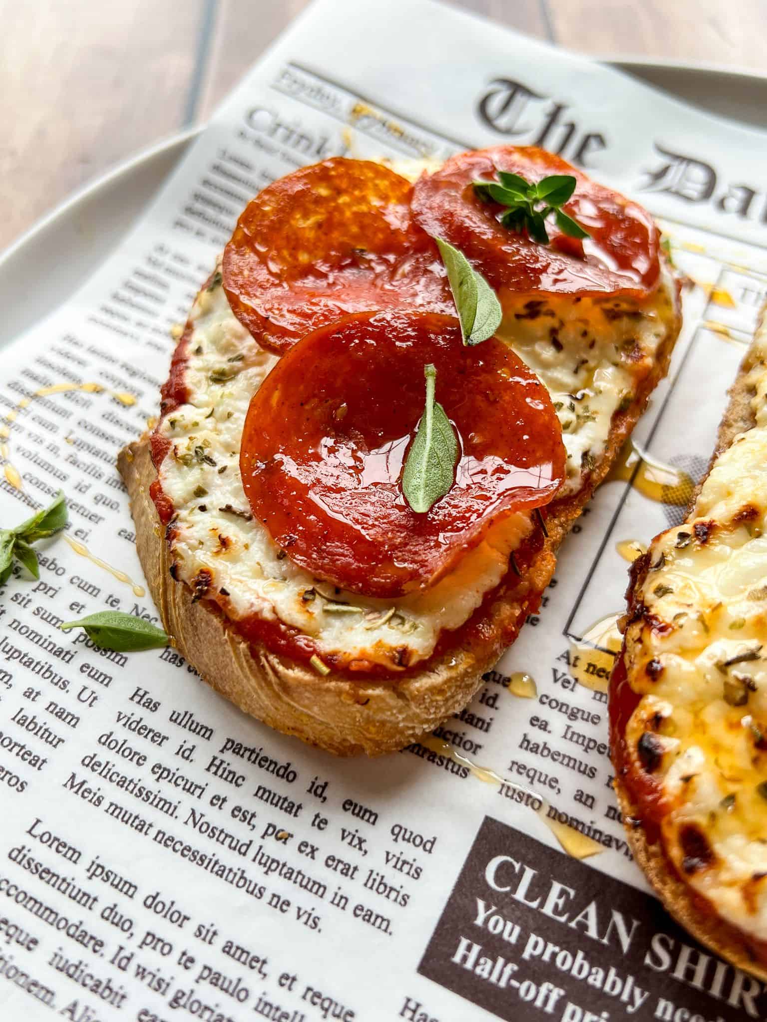 A slice of bread topped with melted cheese, pepperoni slices, and fresh herbs sits on a plate lined with newspaper-style parchment paper. Another similar slice is partially visible beside it.