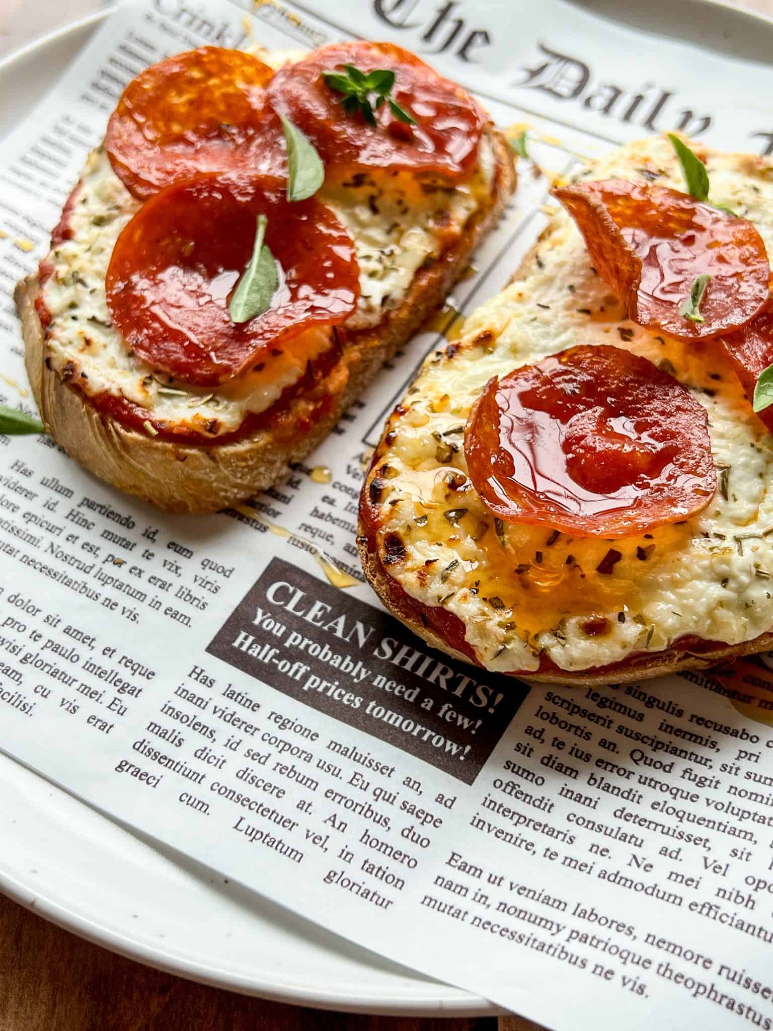Two slices of toasted bread with melted cheese and pepperoni, garnished with fresh herbs, served on a plate lined with newspaper-style parchment.