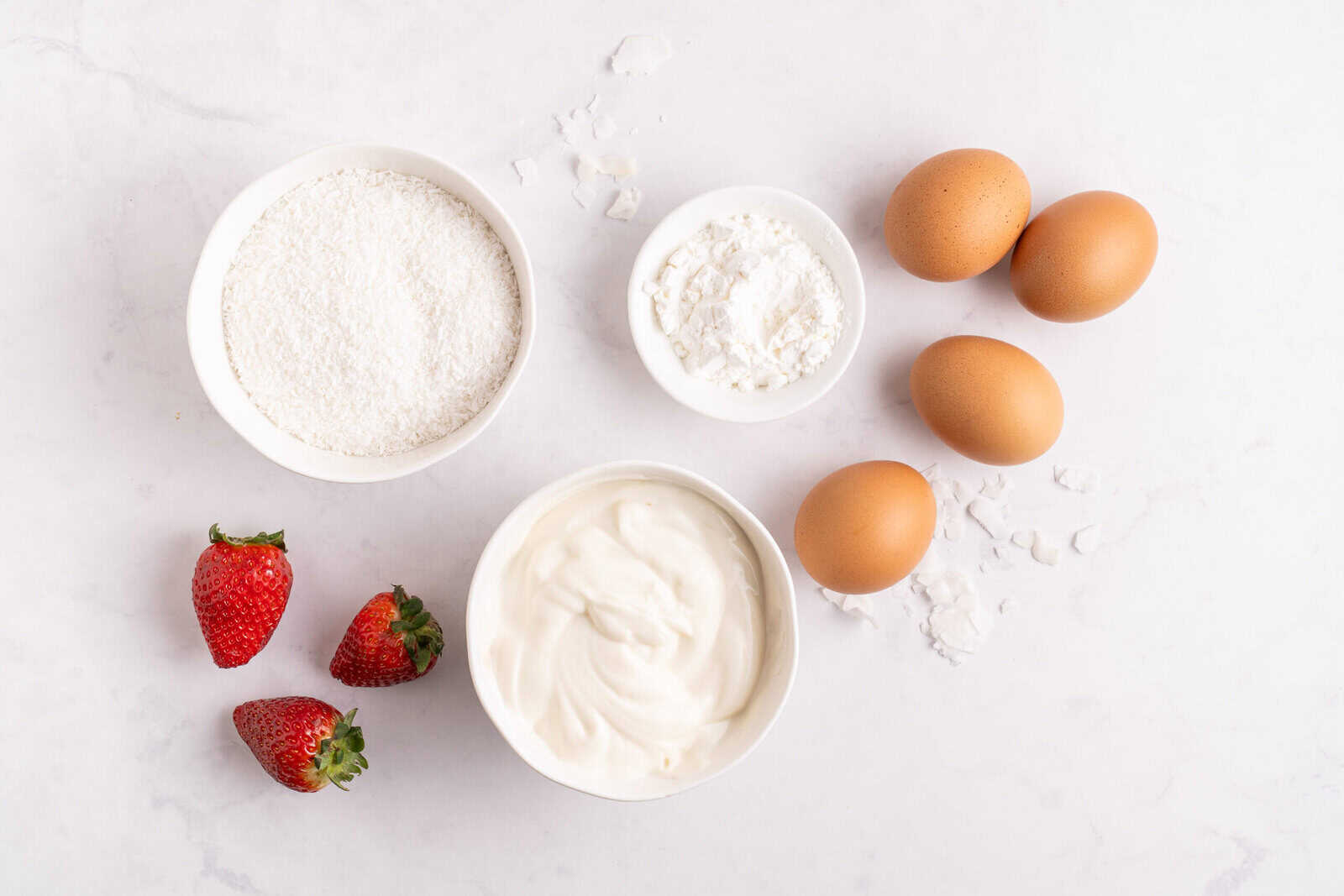 Four brown eggs, two strawberries, a bowl of shredded coconut, a bowl of cornstarch, and a bowl of yogurt are arranged on a white surface. Some loose coconut is scattered around the ingredients.