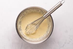 A metal whisk rests inside a stainless steel bowl filled with pale yellow cake batter, placed on a light marble surface.