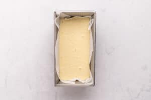 A rectangular loaf pan lined with parchment paper filled with smooth, pale yellow cake batter, sitting on a light marble surface.