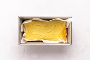 A rectangular loaf of baked bread with a golden top sits in a parchment-lined metal loaf pan on a light surface.