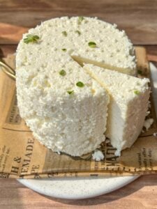 A round, rustic cheese sits on a plate lined with printed paper. A wedge is cut out, revealing its crumbly, white texture. Small green herbs are sprinkled on top. The setup is on a wooden surface in bright natural light.