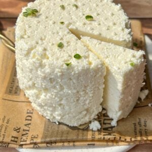 A round, rustic cheese sits on a plate lined with printed paper. A wedge is cut out, revealing its crumbly, white texture. Small green herbs are sprinkled on top. The setup is on a wooden surface in bright natural light.