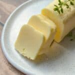 A close-up of three slices of butter on a white speckled plate, with a sprig of fresh thyme placed on the butter. The setting is on a wooden surface.