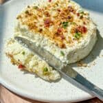 A round, creamy cheese topped with chopped nuts, herbs, and dried fruit sits on a white speckled plate. A knife rests on the plate with a portion of cheese spread beside it.