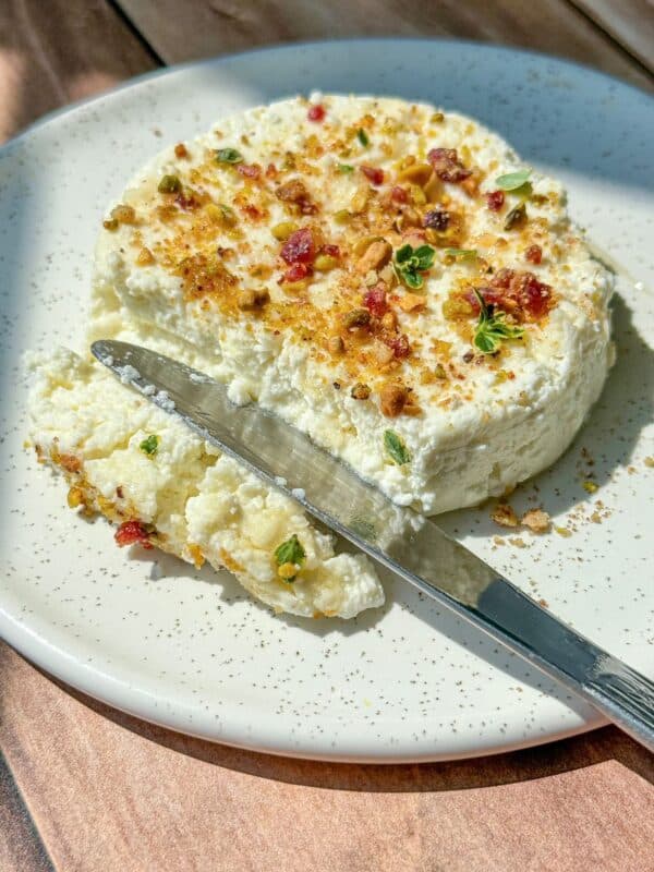 A round, creamy cheese topped with chopped nuts, herbs, and dried fruit sits on a white speckled plate. A knife rests on the plate with a portion of cheese spread beside it.