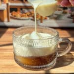 A hand pours creamy milk from a small glass pitcher into a patterned glass mug filled with coffee, set on a wooden surface with sunlight streaming in.