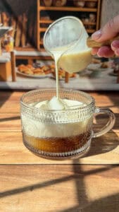 A hand pours creamy milk from a small glass pitcher into a patterned glass mug filled with coffee, set on a wooden surface with sunlight streaming in.